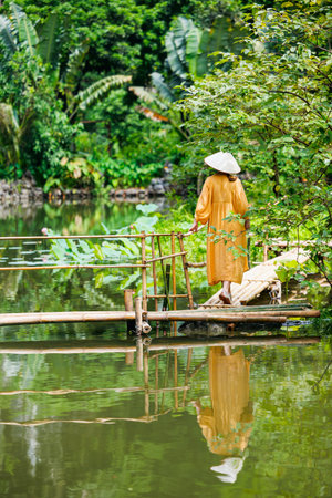 Back view of beautiful woman wearing yellow dress and vietnamese conical in tropical surroundings by lake in Vietnamの写真素材