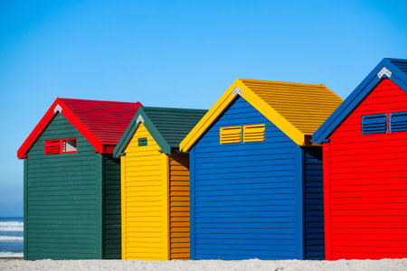 Row of distinctive blue, yellow, red, and green Victorian-style beach changing boxes standing on stilts along Muizenberg's famous sandy shorelineの写真素材