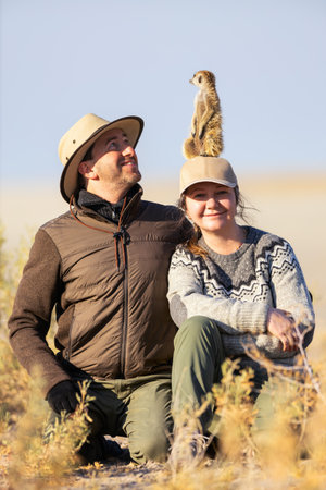 A smiling couple sits on the ground during a safari. A curious meerkat stands perched atop the woman's hat, adding a playful touch to the serene scene.の写真素材
