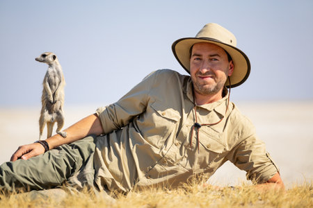 A man in safari attire, reclining on dry grass under the sun, accompanied by a meerkat standing alertly beside him, set against a serene natural backdrop.の写真素材