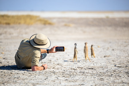 A man in safari gear lying on a dry plain captures a group of standing meerkats with his phone, highlighting a close wildlife encounter in an arid environment.の写真素材