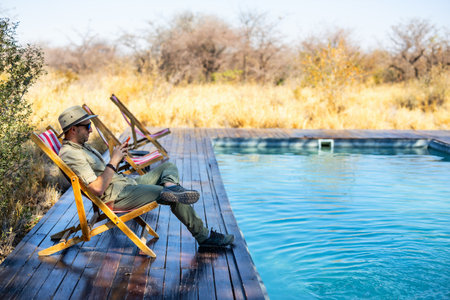 A man in a safari outfit, wearing a hat and sunglasses, reclines on a deck chair beside a pool in the middle of a serene, golden savanna landscape.の写真素材