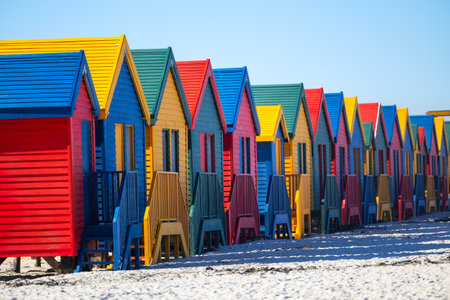 Row of distinctive blue, yellow, red, and green Victorian-style beach changing boxes standing on stilts along Muizenberg's famous sandy shorelineの写真素材