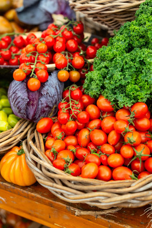 Fresh vine-ripened cherry tomatoes displayed in wicker basket alongside curly kale and purple cabbage at market stand.の写真素材