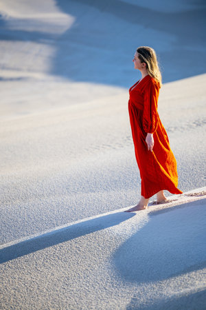 A woman in a vibrant red dress stands barefoot on a serene sand dune, surrounded by soft textures and shadows, illuminated by the gentle afternoon light.の写真素材