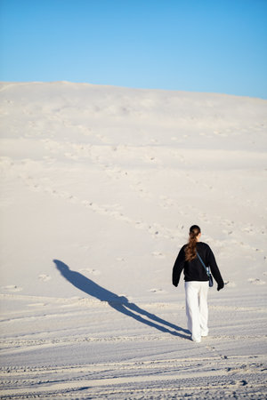 A girl dressed in a black sweater and white pants walks across sandy dunes under clear blue skies. Her long shadow stretches behind her, emphasizing the serene and minimalistic landscape.の写真素材