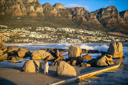 A woman in a flowing dress strolls among the coastal boulders at sunset, framed by Cape Townâs dramatic mountains and ocean waves illuminated by golden light.の写真素材