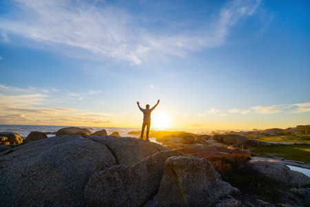 A man stands atop a rocky coastline, arms raised, as the golden sun sets over the ocean, casting a warm glow across the dramatic coastal landscape.の写真素材
