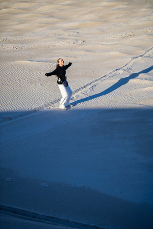 A joyful woman in a black sweater and white pants dances on sand dunes under soft light, her movements casting dynamic shadows on the textured sandy landscape.の写真素材