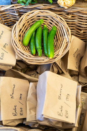 Four fresh green zucchini displayed in rustic wicker basket, surrounded by brown paper bags with handwritten market labels.の写真素材