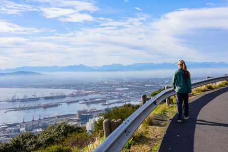 A girl strolls along a scenic pathway overlooking Cape Town's harbor, with cranes, ships, and the city spread below, framed by distant mountains under a bright blue sky.の写真素材