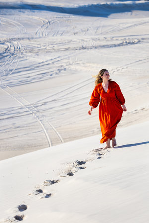 A woman in a vibrant red dress stands barefoot on a serene sand dune, surrounded by soft textures and shadows, illuminated by the gentle afternoon light.の写真素材