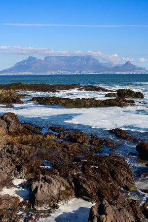 A stunning view of Table Mountain and Lion's Head in Cape Town, seen from a rugged, rocky shoreline with waves crashing and a clear blue sky above.の写真素材