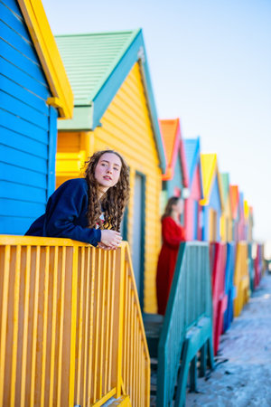 A mother and daughter stand by the vibrant, colorful beach huts at Muizenberg Beach, South Africa, sharing a quiet moment amidst the iconic seaside architecture.の写真素材
