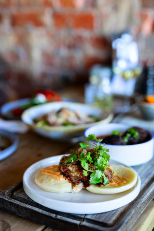 A delicious bao bun dish featuring crispy filling, fresh cilantro, and a creamy sauce, served on a white plate with a rustic wooden table backdrop.の写真素材