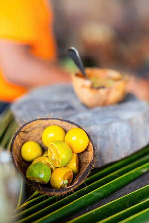 A rustic setup featuring fresh limes in a halved coconut shell, placed on a wooden surface, surrounded by green palm leaves.の写真素材