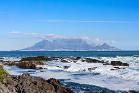 A scenic view of Table Mountain, Lion&#39;s Head, and Cape Town&#39;s skyline in the distance, framed by rocky shores and the vibrant blue ocean with crashing waves in the foreground.の写真素材
