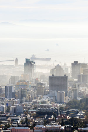 A misty view of Cape Town's skyline, showcasing towering buildings and a bustling harbor with large ships anchored offshore, framed by distant mountains under a hazy sky.の写真素材