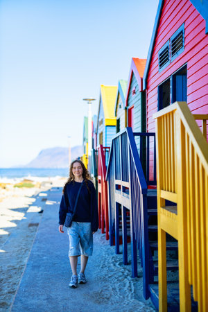 A person stands along the iconic rainbow-colored Victorian beach huts at Muizenberg Beach in Cape Town, with mountains and False Bay in background.の写真素材