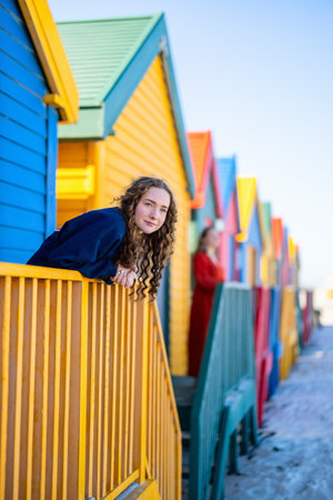 A mother and daughter stand by the vibrant, colorful beach huts at Muizenberg Beach, South Africa, sharing a quiet moment amidst the iconic seaside architecture.の写真素材