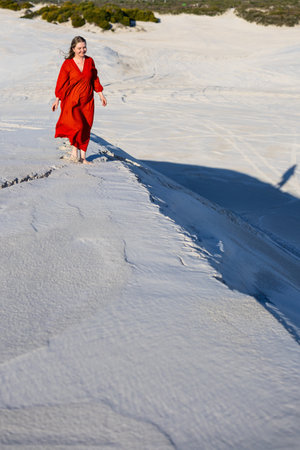 A woman in a flowing red dress walking barefoot along the ridge of a pristine sand dune, surrounded by natural patterns and the warm sunlight of late afternoon.の写真素材
