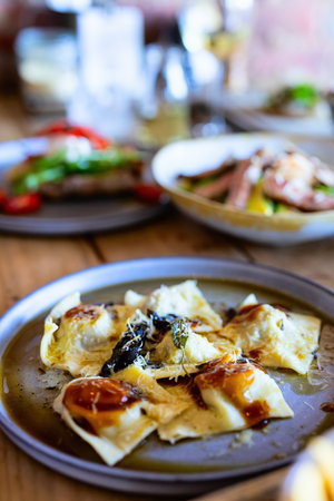 A dish of freshly prepared ravioli topped with parmesan cheese, herbs, and a light sauce, served on a rustic table, with other elegantly plated dishes blurred in the background.の写真素材