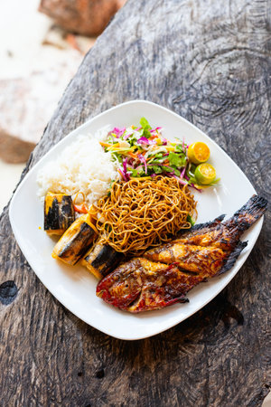 A plate featuring grilled fish, stir-fried noodles, steamed rice, fresh salad, and citrus garnish, served on a rustic wooden table.の写真素材