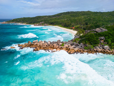 Aerial shot of Grand Anse Beach on La Digue, Seychelles, showcasing turquoise waters, crashing waves, golden sand, lush greenery, and granite rock formations.の写真素材