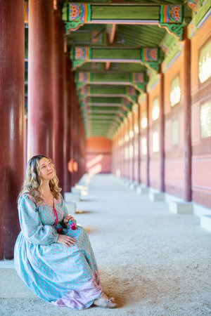 A tourist in a flowing, patterned dress sits peacefully on the floor of a vibrant, traditional Seoul temple, surrounded by beautifully carved wooden pillars and intricate architecture.の写真素材