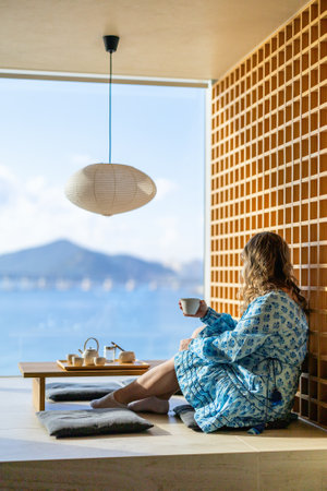 A woman sits in a serene apartment, enjoying green tea, with a breathtaking view of the ocean and distant mountains through a large window.の写真素材