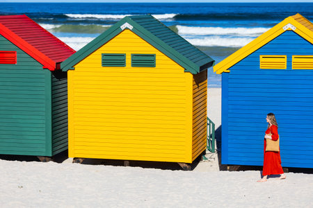 Vibrant beach houses in green, yellow, and blue line the white sandy beach while a woman in a red dress carries a straw bag alongside themの写真素材