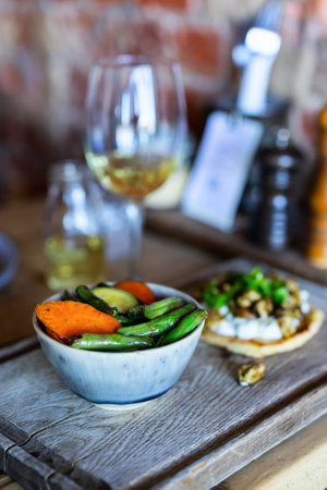 A blue ceramic bowl filled with roasted sweet potatoes and snap peas, accompanied by white wine glasses and an artisanal appetizer plate.の写真素材
