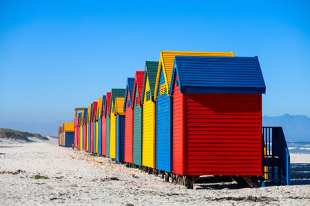 Row of distinctive blue, yellow, red, and green Victorian-style beach changing boxes standing on stilts along Muizenberg's famous sandy shorelineの写真素材