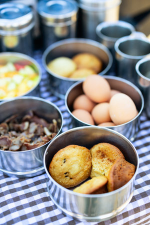 An assortment of breakfast items, including eggs, muffins, bacon, and fruit, served in shiny metal containers on a checkered tablecloth, showcasing a simple outdoor dining setup.の写真素材