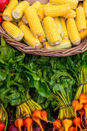 A vibrant display of fresh vegetables at a farmers market, featuring yellow corn on the cob in a wicker basket, alongside bundles of leafy greens and golden beetsの写真素材