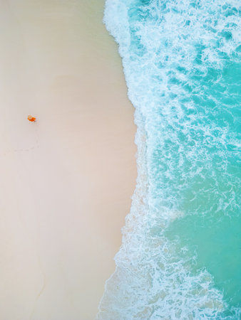 Aerial shot of a person walking along a white sandy beach, leaving footprints near the turquoise ocean waves in a serene and isolated coastal sceneの写真素材
