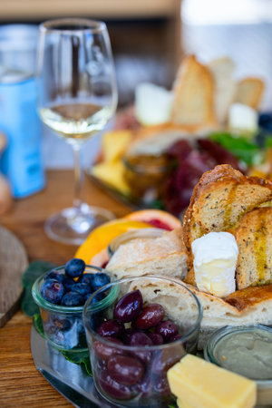A beautifully arranged tapas spread featuring sourdough bread, creamy cheese, olives, blueberries, and other fresh ingredients, accompanied by a glass of white wine on a wooden table.の写真素材