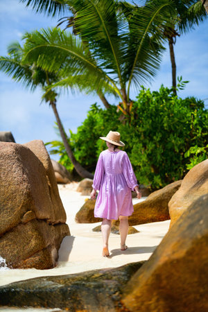 A woman in a lilac dress and sun hat walking barefoot on a tropical beach, surrounded by large rocks, palm trees, and lush greeneryの写真素材