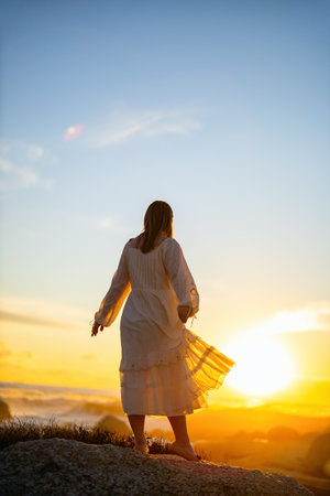 A woman in a flowing white dress stands on a rocky hill, gazing at a vibrant sunset. Her dress catches the light as the sun illuminates the horizonの写真素材