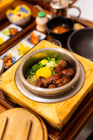 A beautifully presented Korean dish featuring seared beef, raw egg yolks, chopped green onions, sesame seeds, and wasabi served in a heated stone bowl with various side dishesの写真素材