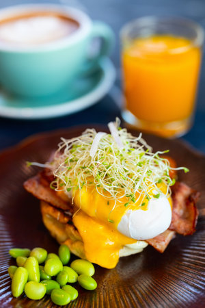 A gourmet serving of Eggs Benedict topped with fresh sprouts and creamy hollandaise sauce, accompanied by edamame, with a cappuccino and orange juice in the backgroundの写真素材