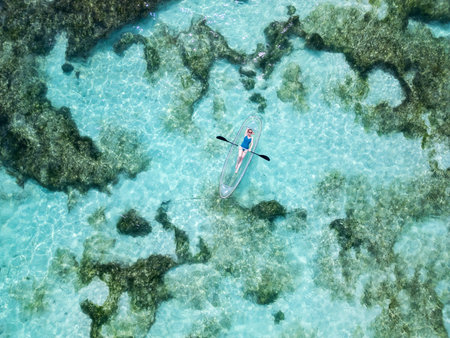 Aerial view of a woman reclining in a transparent kayak floating over shallow, clear turquoise waters with visible underwater coral formations in Seychellesの写真素材