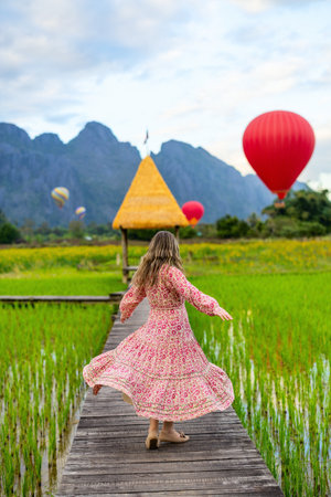 In Vang Vieng, Laos, a woman joyfully twirls on a wooden walkway through a rice paddy, with vibrant hot air balloons and a thatched pavilion in the backgroundの写真素材