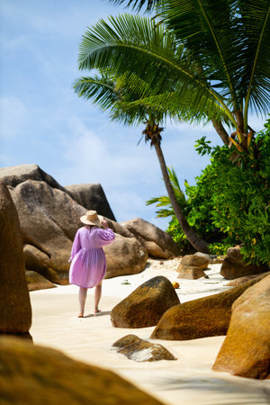 A woman in a lilac dress and sun hat walking barefoot on a tropical beach, surrounded by large rocks, palm trees, and lush greeneryの写真素材