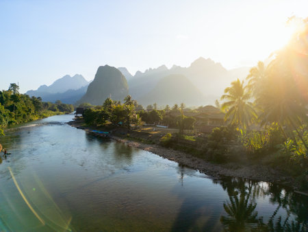 An aerial image of Vang Vieng, Laos, showcasing a serene river winding through the landscape, with lush greenery, palm trees, and surrounding karst mountains at sunsetの写真素材