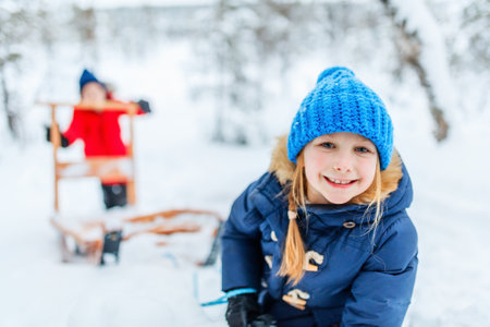 A cheerful child wearing a blue knit hat and winter jacket smiles in a snowy forest in Finnish Lapland, with a sled and another child in the backgroundの写真素材