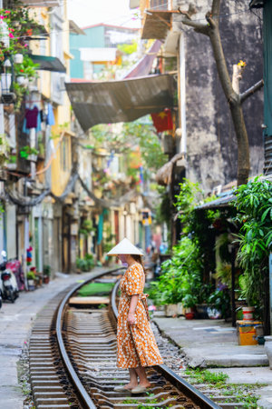 A woman in a floral dress and traditional Vietnamese conical hat stands on the tracks of Hanoi's famous Train Street, surrounded by vibrant, narrow buildingsの写真素材