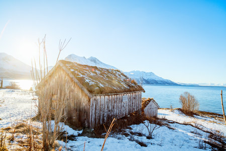 A rustic wooden cabin with a sod roof sits by the seashore in Norway, surrounded by snow-covered terrain and backed by distant mountains under a clear skyの写真素材