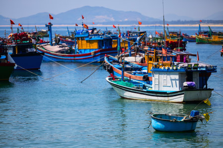 A vibrant scene of colorful fishing boats anchored in calm waters near a coastal area in Vietnam, with mountains in the background and national flags flyingの写真素材