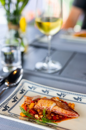 A dish featuring grilled squid served on a bed of diced vegetables in a rich sauce, garnished with a sprig of fresh dill. A glass of white wine is in the backgroundの写真素材